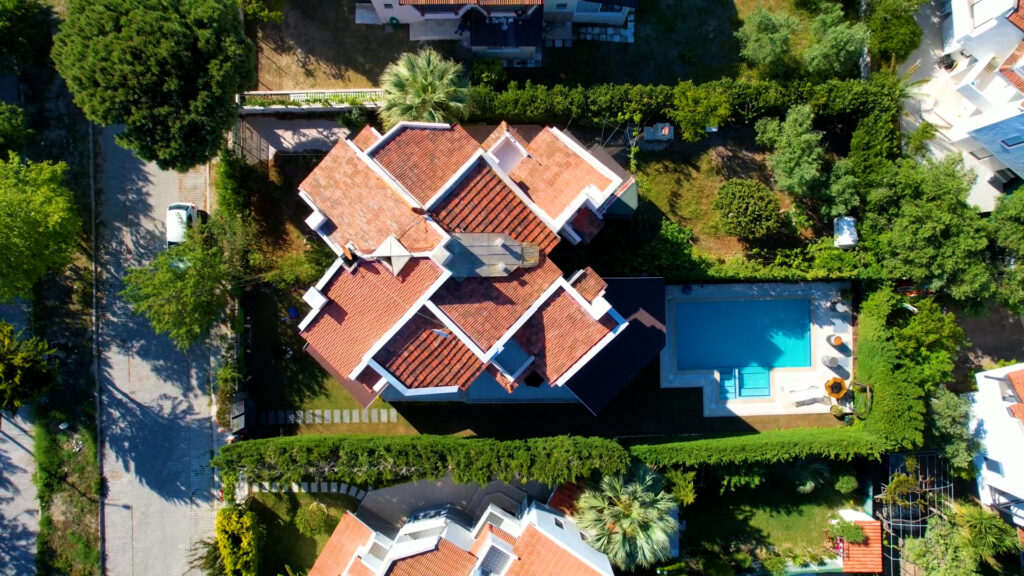 Çeşme Sakarya Mahallesi'nde yer alan havuzlu bir ikiz villanın kuş bakışı (drone) çekimi. Kiremit çatılı villa, yüksek yeşil çitlerle çevrili geniş bir bahçeye sahip. Bahçenin bir bölümünde turkuaz renkli bir yüzme havuzu, güneşlenme alanı ve düzenli peyzaj öğeleri görülüyor.