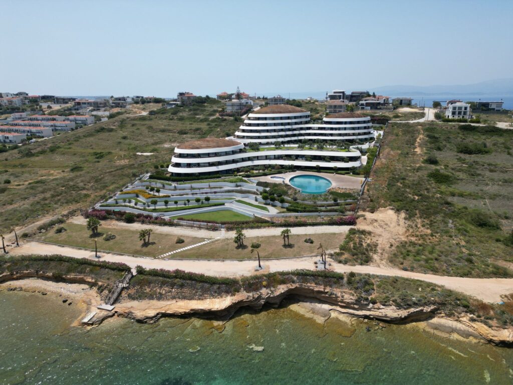 Çeşme sahil şeridinde, denize sıfır konumda yer alan, modern ve kıvrımlı mimariye sahip bir rezidans binası. Binanın önünde kademeli peyzaj alanları, bir yüzme havuzu ve kıyı şeridinde berrak bir deniz görülüyor.