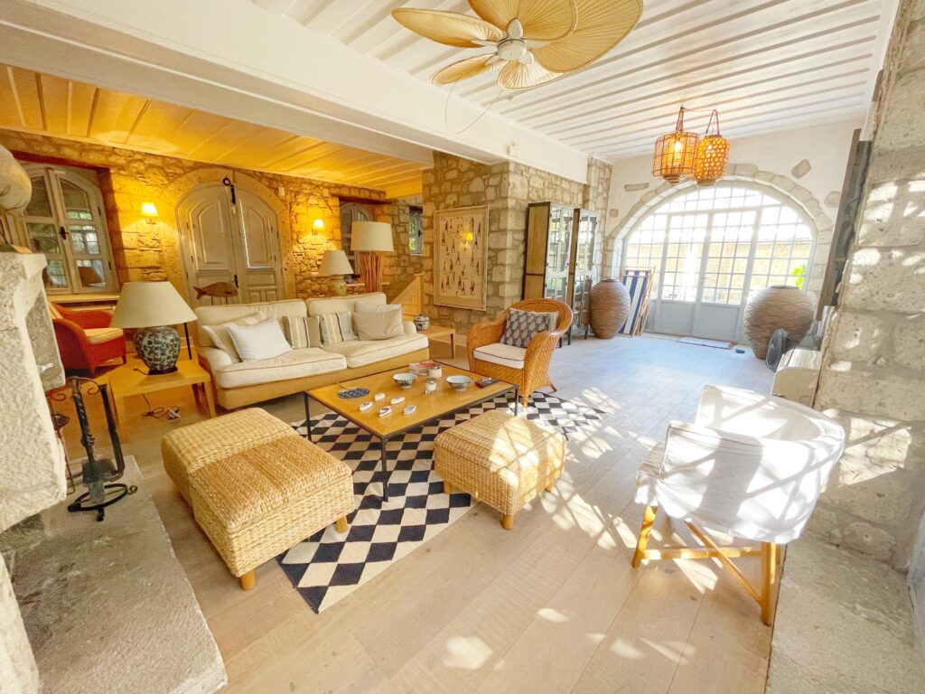 Alaçatı taş mimarisine sahip, yüksek tavanlı, rustik ve modern dekorasyonun harmanlandığı geniş bir oturma odası. Odada beyaz koltuklar, hasır puf ve koltuklar, damalı bir halı ve büyük kemerli bir cam kapı bulunuyor.
