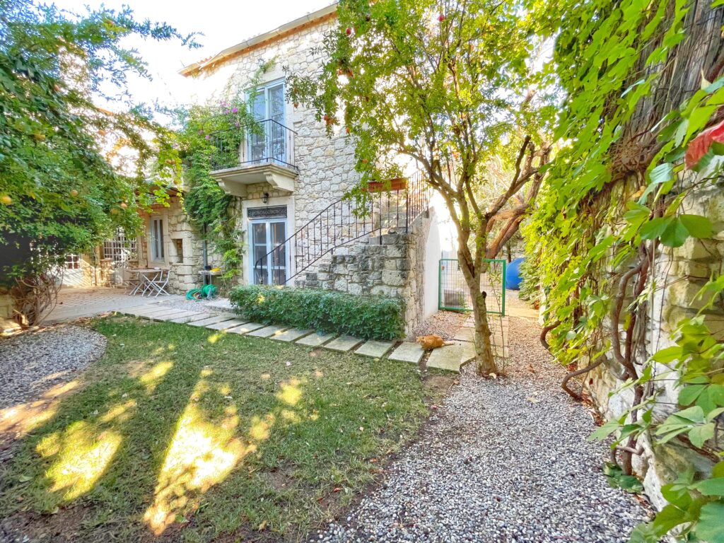 Alaçatı'da geleneksel taş mimarisiyle inşa edilmiş iki katlı bir evin bahçesi. Taş duvarlara sarılmış sarmaşıklar, meyve yüklü nar ağaçları, dışarıdan üst kata çıkan taş bir merdiven ve çimenlerin üzerinde güneşlenen turuncu bir kedi görülüyor. Bahçede taş yürüyüş yolu ve beyaz ferforje bir masa takımı bulunuyor.