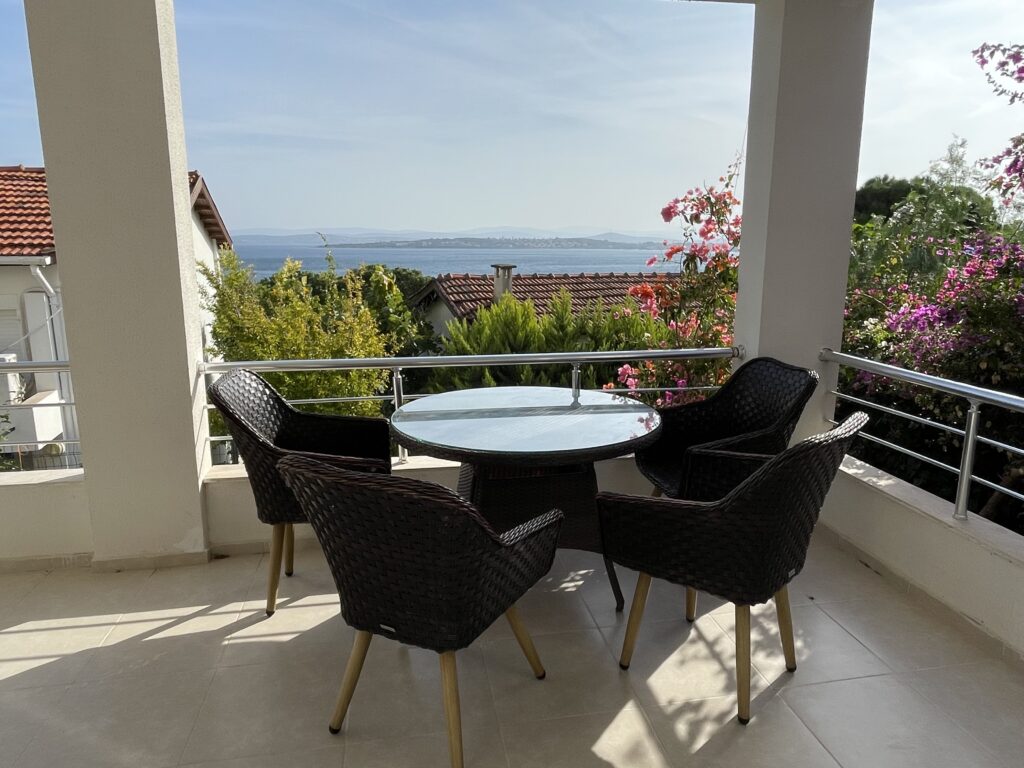 Çeşme Sakarya Mahallesi'nde denize nazır bir villanın geniş balkonundan görünüm. Balkonda modern hasır sandalyeler ve cam tablalı bir yemek masası bulunuyor. Manzarada turkuaz Ege denizi, karşı kıyıdaki adalar, begonviller ve komşu evlerin kiremit çatıları görülüyor.