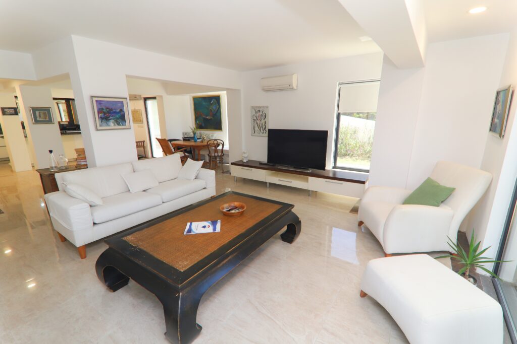 Dalyan Marina yakınındaki villanın geniş salonuna farklı bir bakış. Beyaz modern koltuklar, geniş siyah ahşap orta sehpa ve parlak mermer zeminler ön planda. Arka planda ise mutfağa ve yemek masasına açılan açık plan yerleşim, televizyon ve modern tablolar görülüyor.