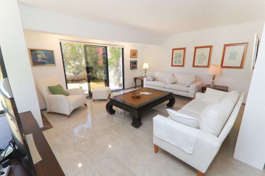 Çeşme Dalyan Marina yakınında, modern dekorasyonlu bir villanın aydınlık oturma odası. Odada beyaz geniş koltuklar, koyu ahşap klasik bir orta sehpa, parlak mermer zeminler ve bahçeye açılan büyük sürgülü cam kapılar bulunuyor. Duvarda tablolar ve şık masa lambaları yer alıyor.