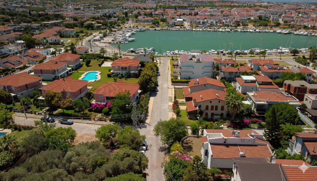 Çeşme Dalyan Mahallesi'ndeki bir villanın balkonundan çekilen, Dalyan Marina ve Ege Denizi manzaralı drone fotoğrafı. Ön planda yeşil bahçeler, begonviller ve kiremit çatılı evler, arka planda ise marinadaki yatlar, tekneler ve masmavi deniz görülüyor.